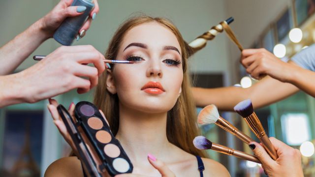 Portrait of the charming young woman who is sitting indoors and makeup artist doing makeup her and hairdresser doing the hairdo her