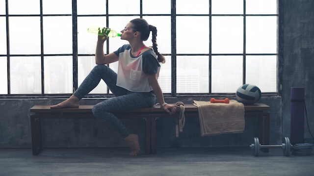Fit woman in profile sitting on bench in loft gym drinking water