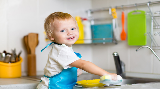 Toddler child washing dishes in kitchen. Little boy having fun with helping to his mother with housework.